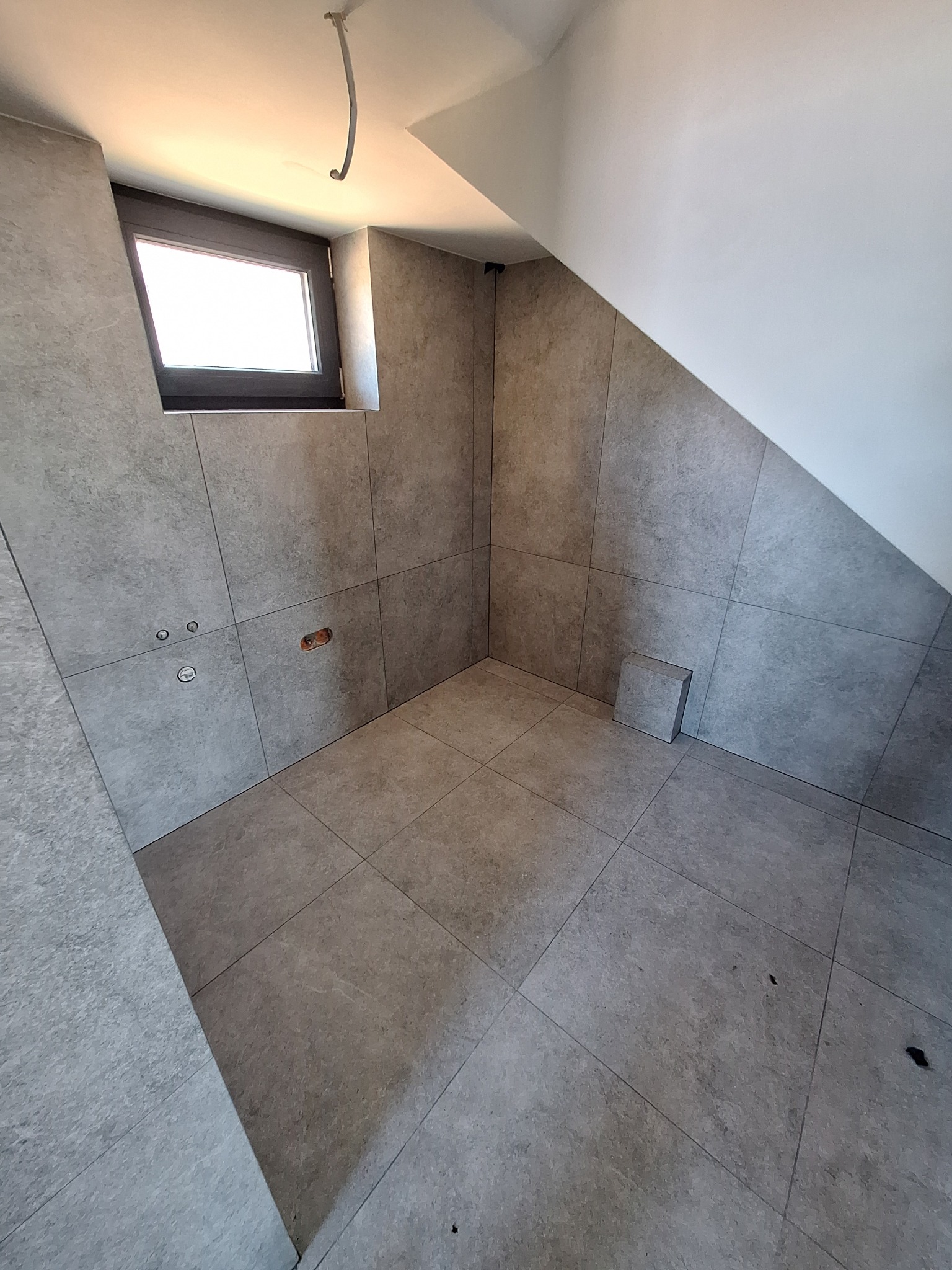 Wykończona łazienka na poddaszu ze skośnym sufitem, wyłożona szarymi, matowymi płytkami. Widoczne okno i wystające rury instalacyjne.