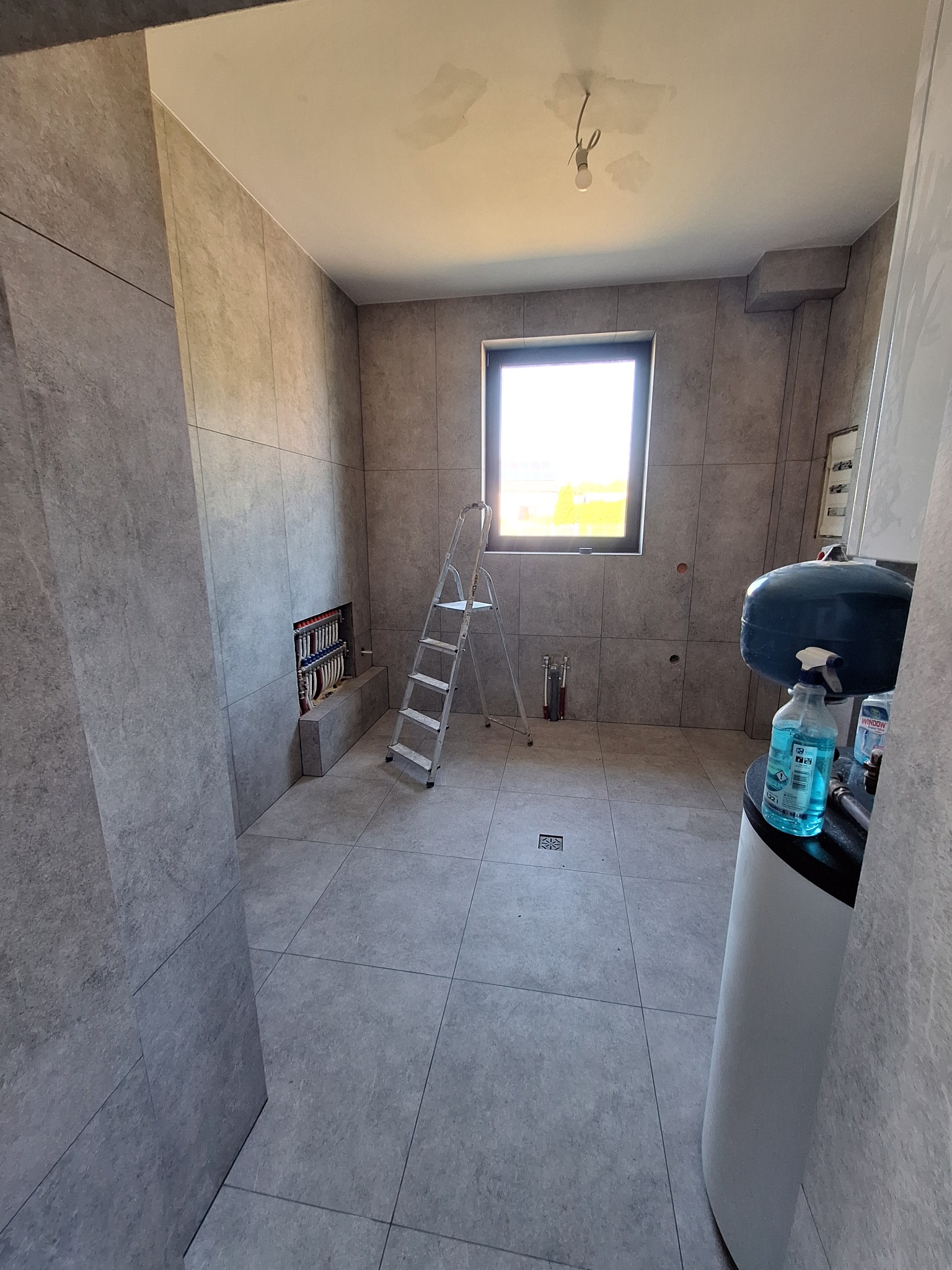 Szara łazienka w trakcie prac wykończeniowych: płytki na ścianach i podłodze, drabina, instalacja grzewcza w ścianie, okno z widokiem na zewnątrz.