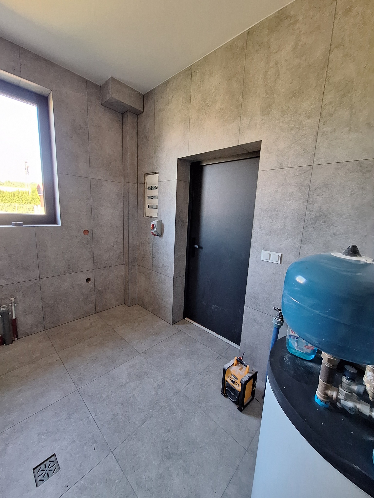 Wykończona łazienka w szarych płytkach z widocznym oknem, drzwiami, bojlerem i narzędziami. Ujęcie z bliska, prezentujące detale wykończenia.