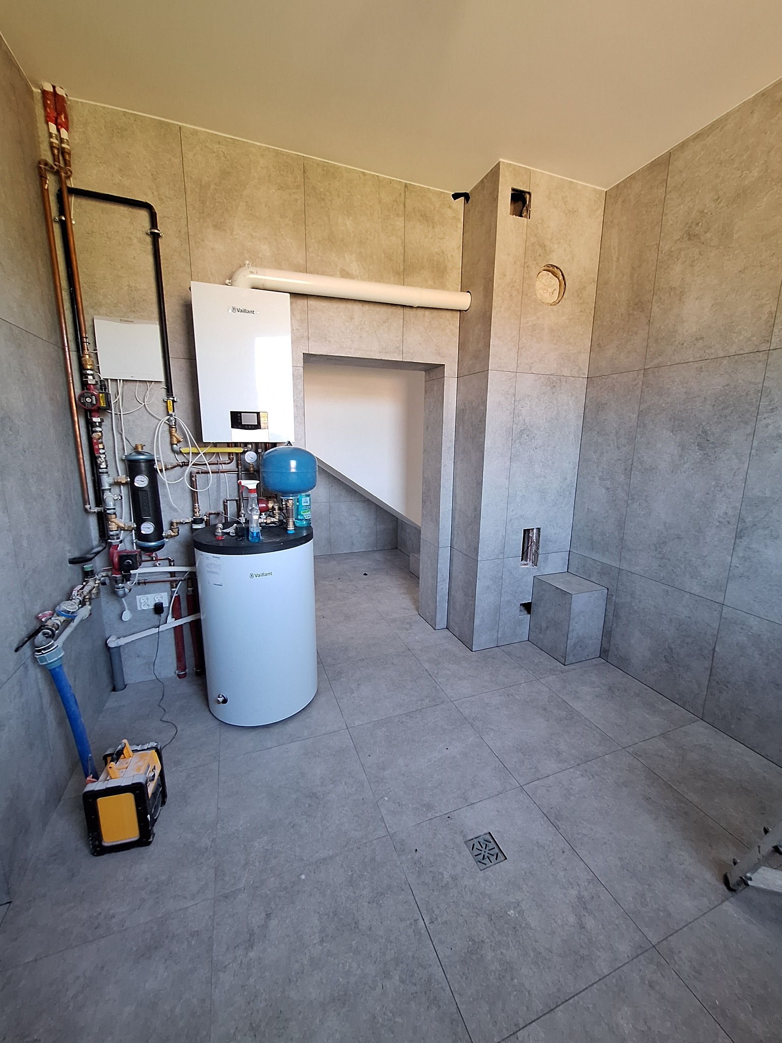 Szara łazienka z zamontowanym piecem gazowym Vaillant, instalacją hydrauliczną i wentylacją. Widoczny odpływ podłogowy i skrzynka narzędziowa.