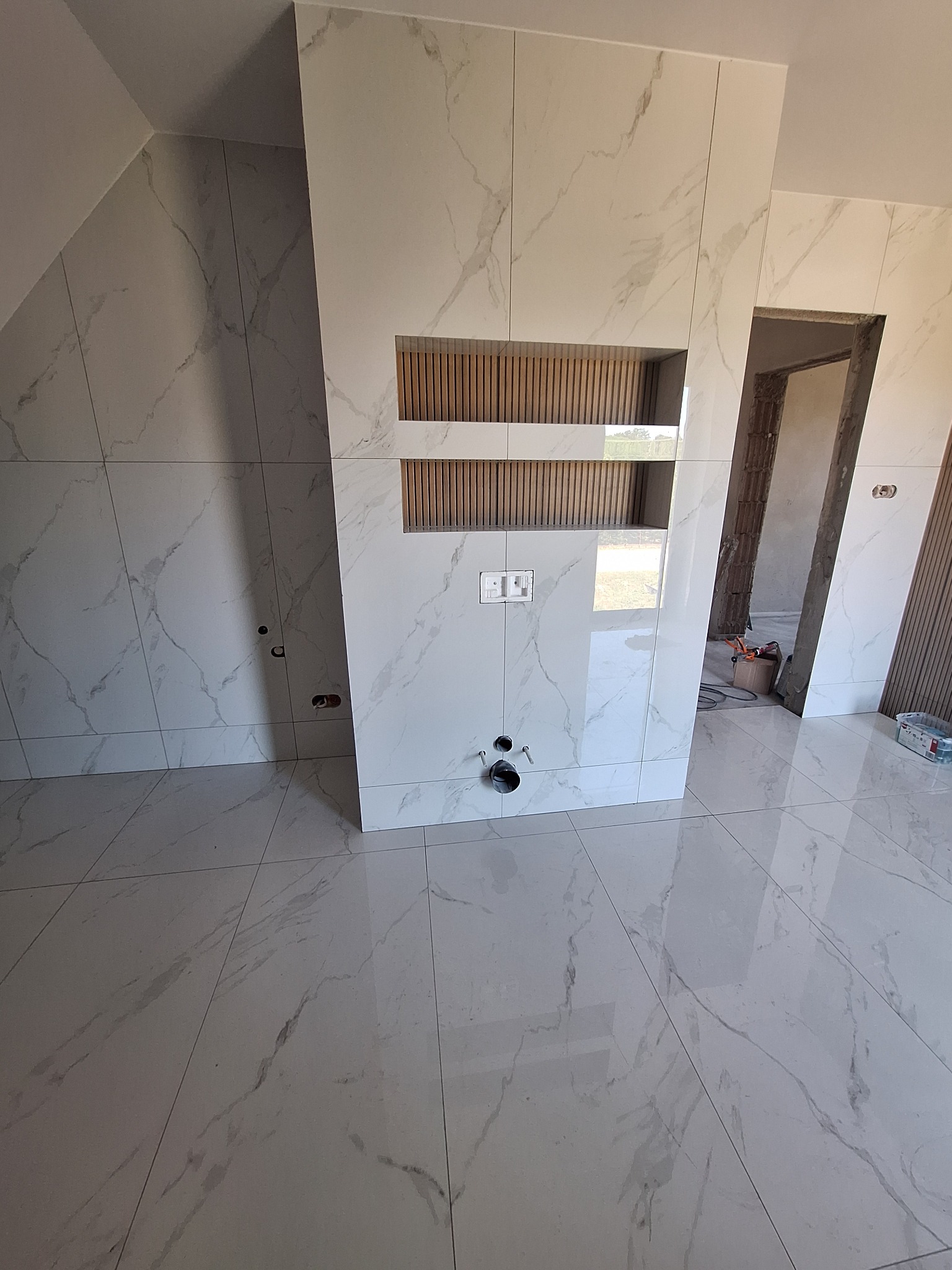 Wykończona łazienka z białymi płytkami imitującymi marmur, drewniane akcenty w niszach. Widoczne przyłącza hydrauliczne i elektryczne. Otwór drzwiowy w tle.