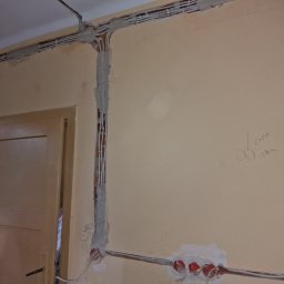 Smart Electrics Przemysław Sowa - Ściana w trakcie remontu z widocznymi przewodami elektrycznymi w bruzdach, częściowo zamaskowanymi gipsem, oraz puszkami instalacyjnymi z czerwonymi elementami w dolnej części ściany, obok...