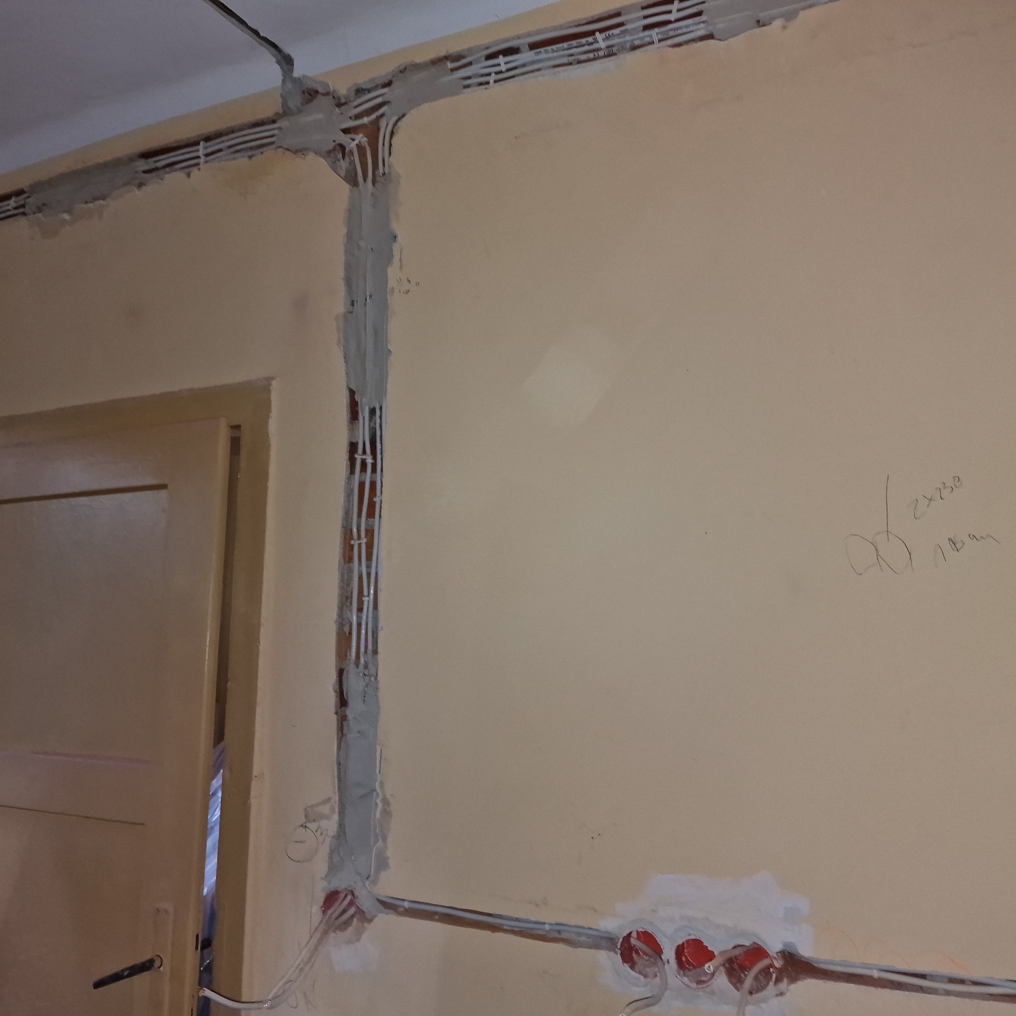 Ściana w trakcie remontu z widocznymi przewodami elektrycznymi w bruzdach, częściowo zamaskowanymi gipsem, oraz puszkami instalacyjnymi z czerwonymi elementami w dolnej części ściany, obok...