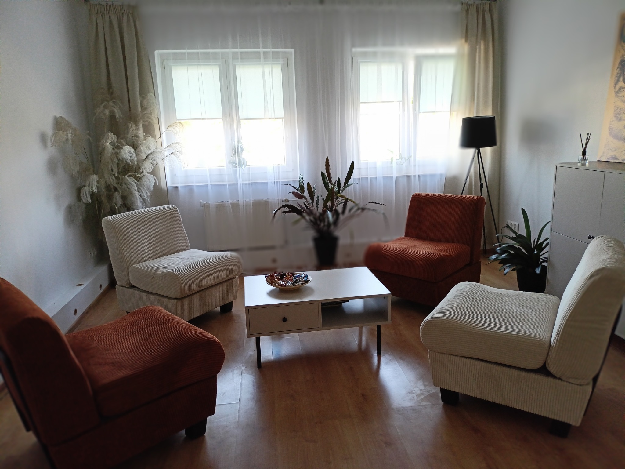 Jasny gabinet z czterema fotelami (dwoma beżowymi i dwoma pomarańczowymi) ustawionymi wokół białego stolika kawowego z misą cukierków, widok na okno z białymi zasłonami i roślinami.