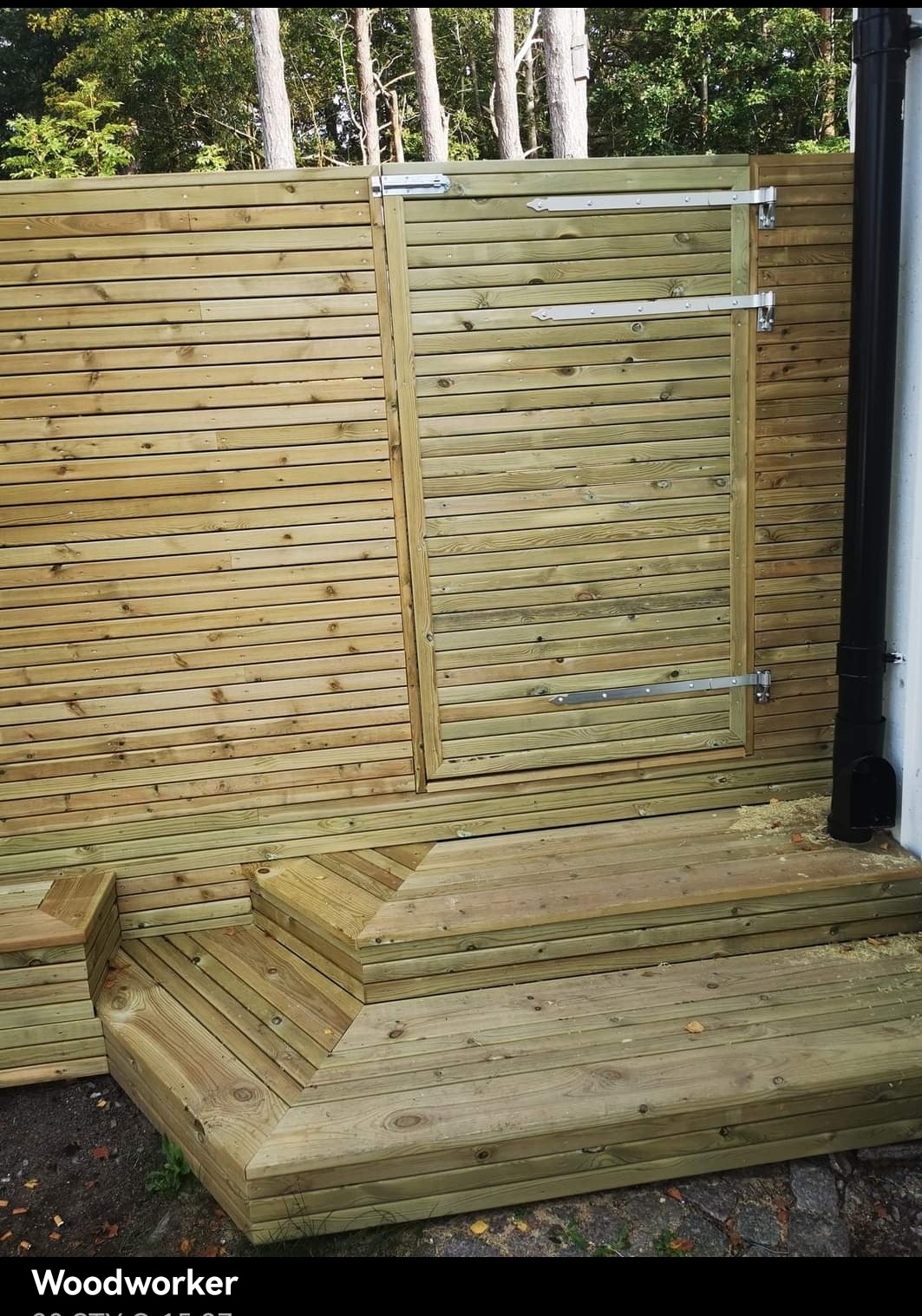 Drewniana konstrukcja z furtką i schodami, wykonana z desek w naturalnym odcieniu drewna, widoczna na tle zieleni.