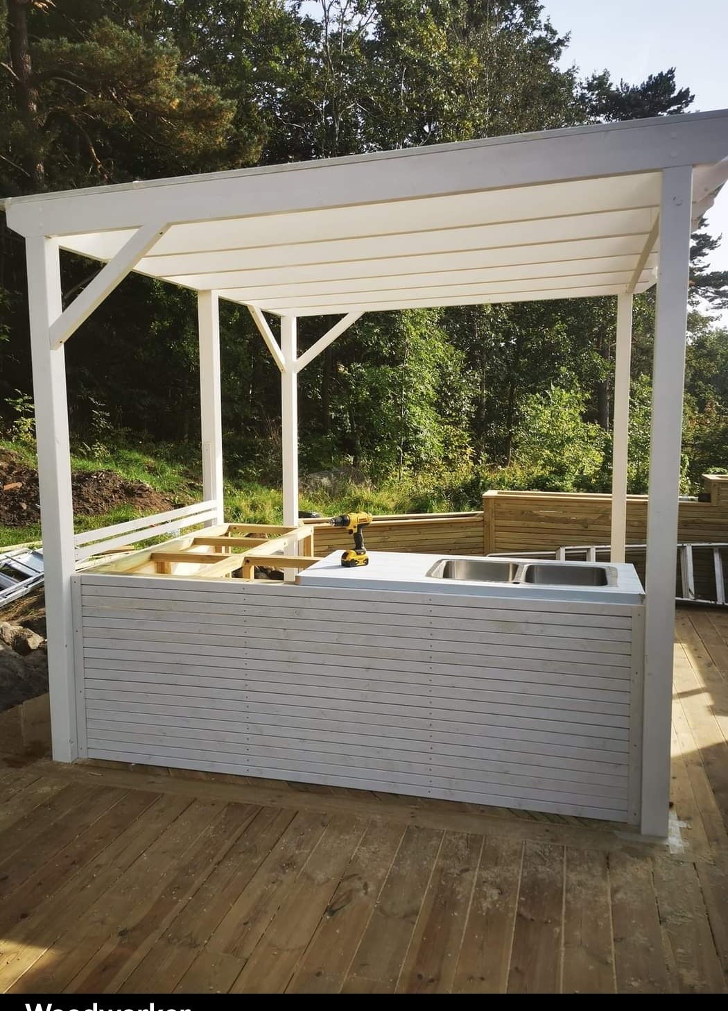Biała, drewniana altana w trakcie budowy, widoczne elementy konstrukcyjne, narzędzia i wbudowany zlew ze stali nierdzewnej na tle zieleni.