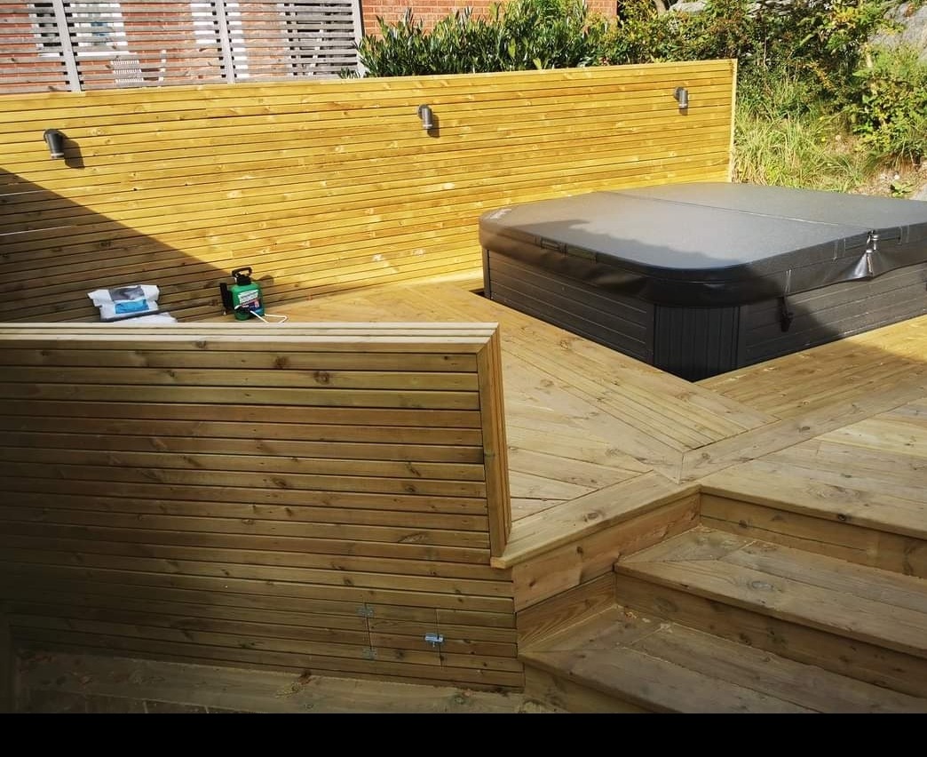 Drewniany taras z jacuzzi i zabudową z poziomych desek, oświetlenie punktowe, widoczne schody i akcesoria do czyszczenia.