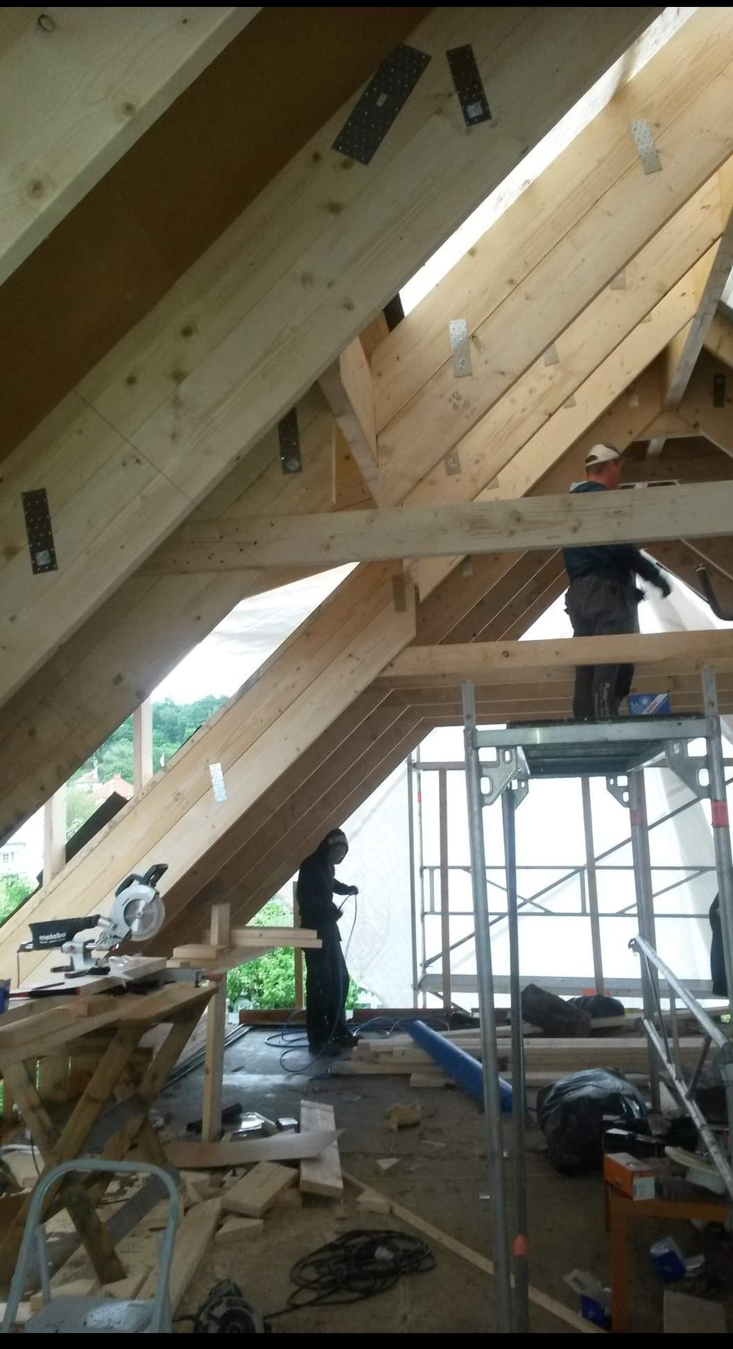 Wnętrze budowanego domu szkieletowego: widoczne drewniane belki konstrukcyjne, pracownicy na rusztowaniu oraz narzędzia, w tle zieleń.