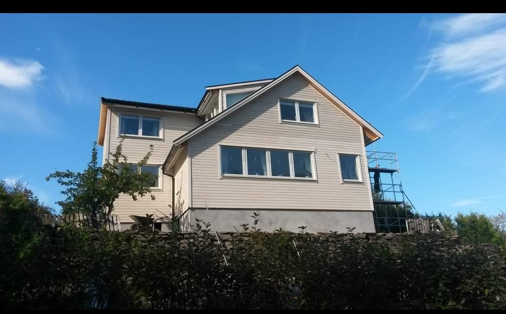 Dwukondygnacyjny dom z beżową elewacją sidingową, widoczny zza kamiennego murku i zielonych krzewów, z rusztowaniem z tyłu i błękitnym niebem w tle.