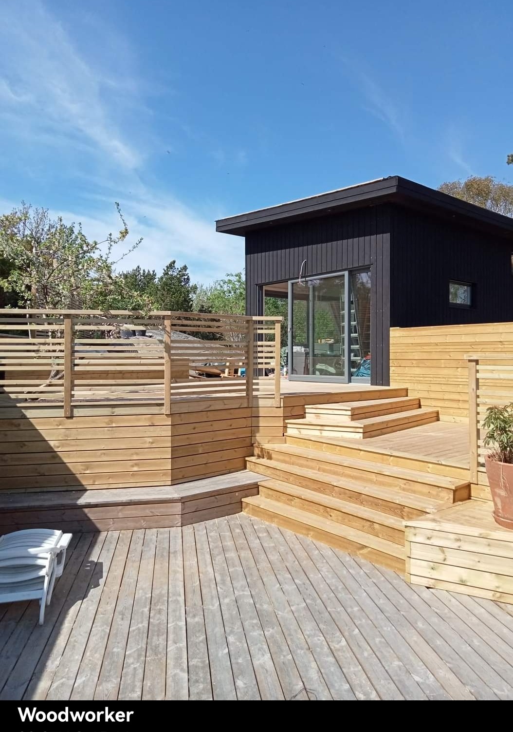 Nowoczesny, czarny budynek z przesuwnymi szklanymi drzwiami, otoczony drewnianym tarasem wielopoziomowym z barierkami i schodami, widok w słoneczny dzień.