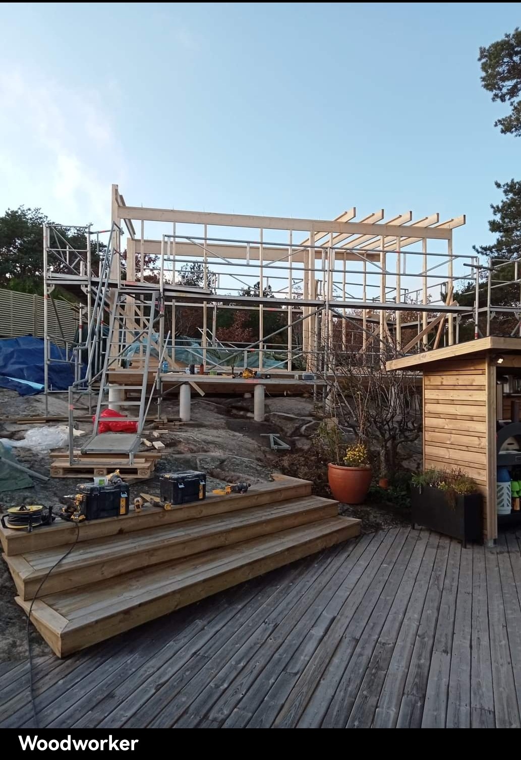 Konstrukcja szkieletowa domu na skalistym terenie, widoczne rusztowania, narzędzia i drewniane schody prowadzące na taras.