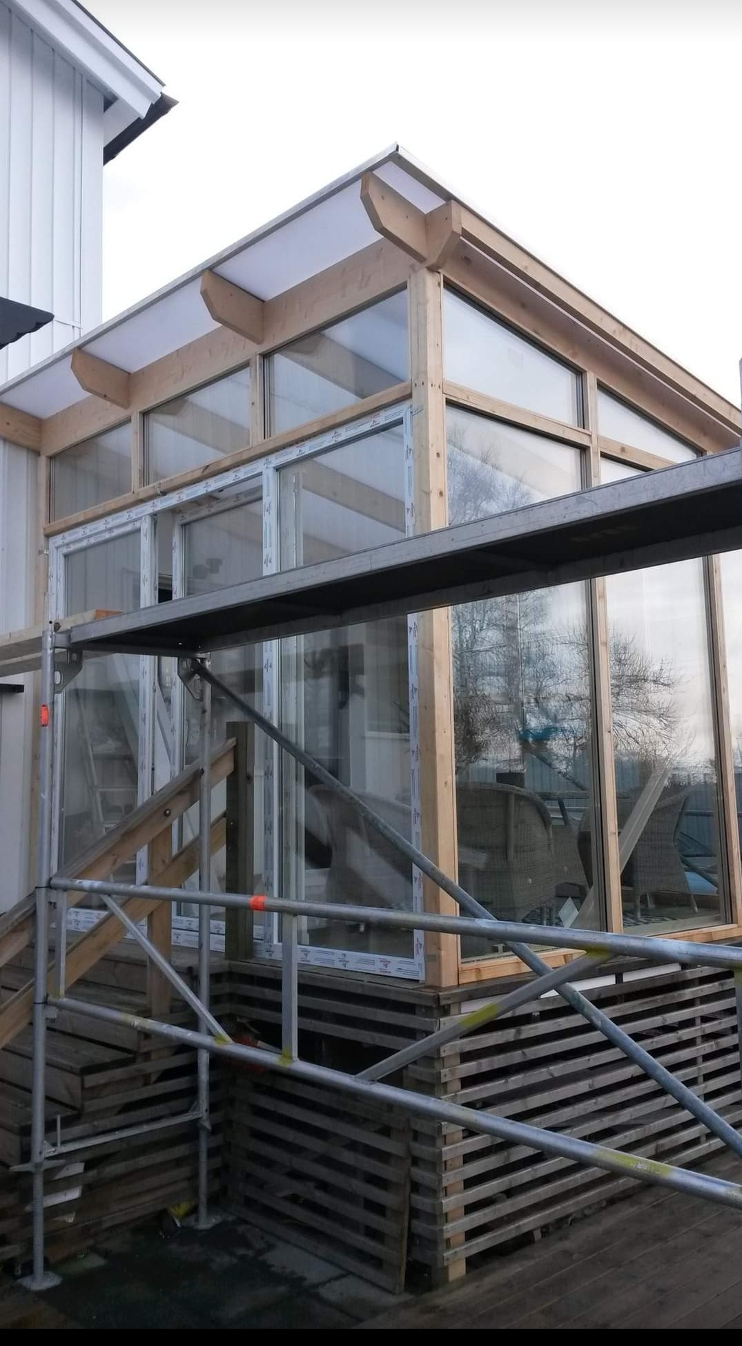 Konstrukcja przeszklonej werandy z drewnianym szkieletem, widoczna z rusztowania, w tle dom z białą elewacją.