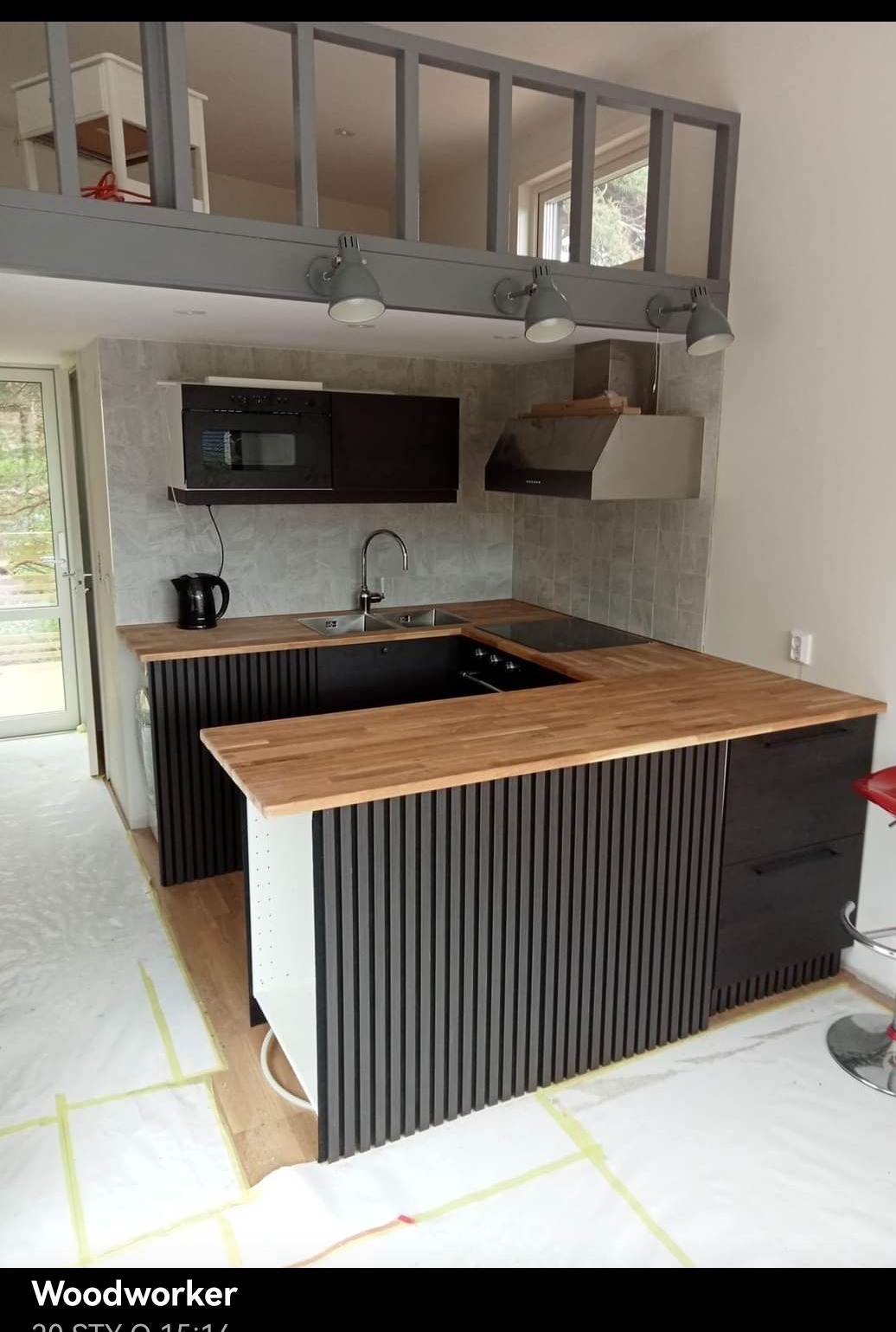 Nowoczesna kuchnia z drewnianym blatem, czarnymi szafkami z pionowymi listwami i antresolą z szarej stali. Widoczne cztery szare lampy punktowe, czajnik elektryczny i okap.