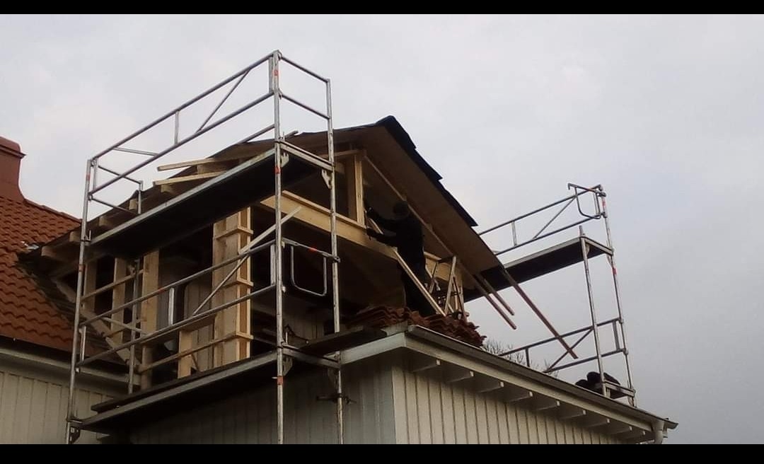 Prace na dachu domu podczas rozbudowy poddasza, widoczne rusztowania i konstrukcja szkieletowa, częściowo pokryta płytami, pracownik w ciemnym ubraniu na rusztowaniu.