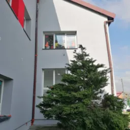 Szary budynek z nową elewacją, brązowe rynny, okna z dekoracjami i zielony krzew iglasty na pierwszym planie.