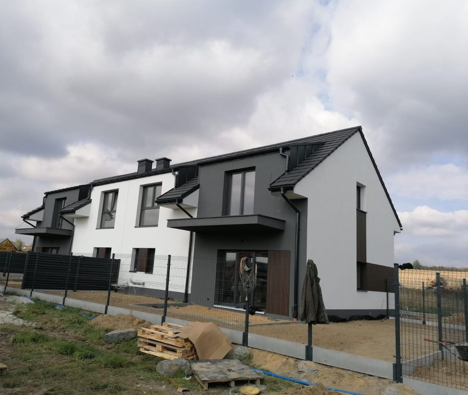 Nowoczesne szeregowe domy z biało-szarą elewacją i ciemną stolarką okienną, ogrodzone siatką, z widocznymi materiałami budowlanymi na niezagospodarowanym terenie.