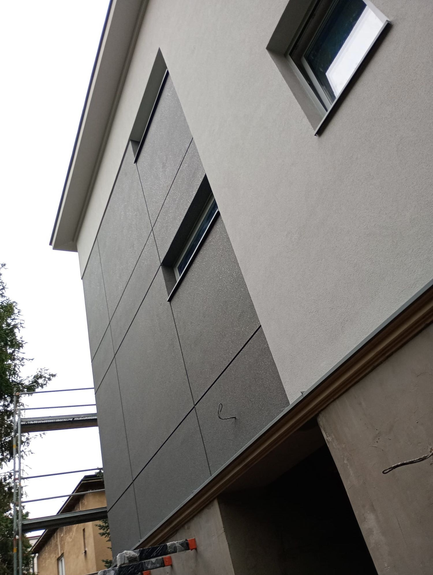 Fragment elewacji budynku z szarą, podzieloną na kwadraty powierzchnią, widoczne okna i rusztowanie w tle.