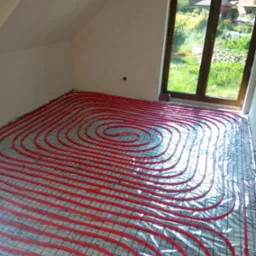 Czerwone rury ogrzewania podłogowego ułożone spiralnie na folii z siatką w jasnym pokoju na poddaszu z oknem.