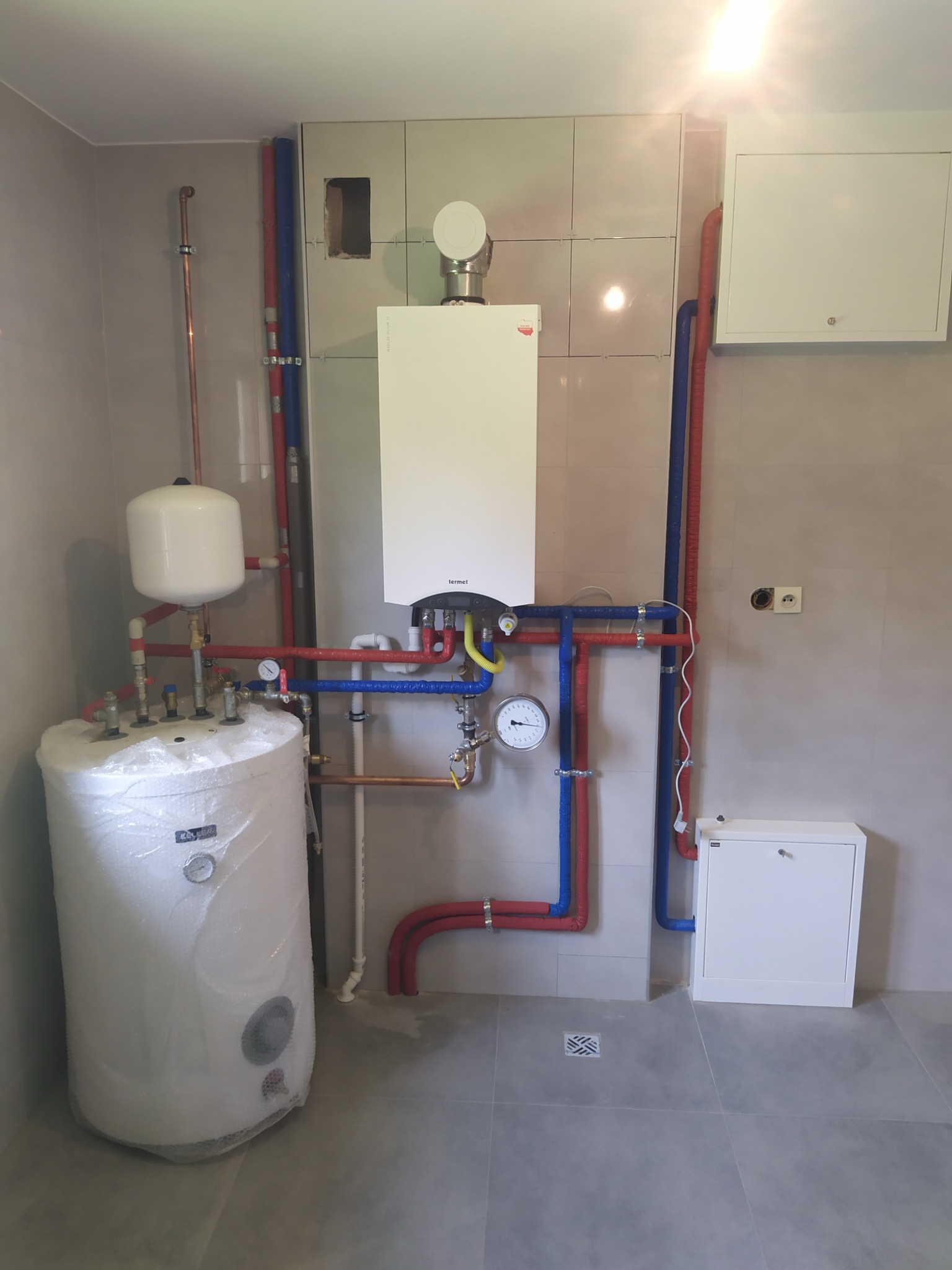 Nowoczesny piec gazowy wiszący Termet, podgrzewacz wody w folii ochronnej, instalacja z rur miedzianych i plastikowych w kolorach czerwonym i niebieskim na tle jasnych płytek ceramicznych i szarej...