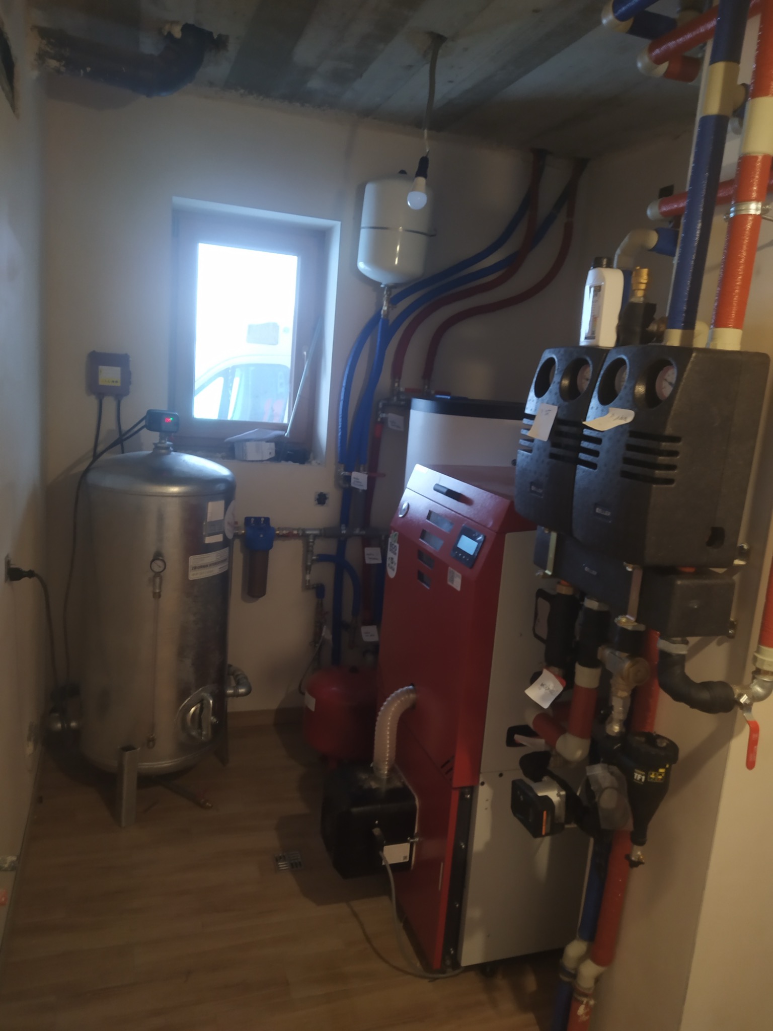 Kompletna kotłownia z piecem CO, zasobnikiem CWU i rozbudowaną instalacją hydrauliczną z rur PEX w nowym budynku w trakcie wykańczania, widoczne liczne zawory, filtry i pompy obiegowe.