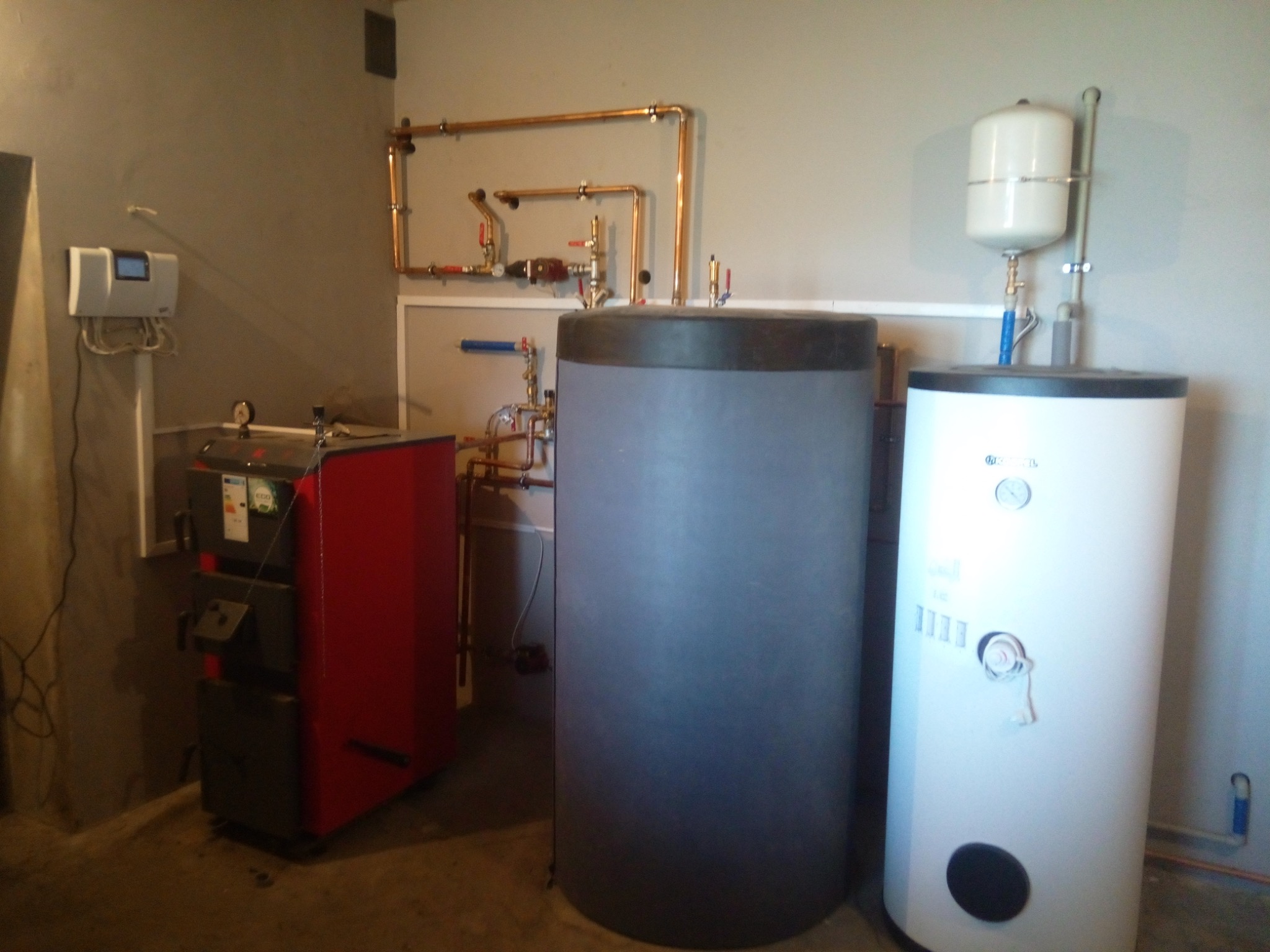 Kotłownia z kotłem na paliwo stałe, dwoma zbiornikami akumulacyjnymi i miedzianą instalacją centralnego ogrzewania na szarej ścianie.