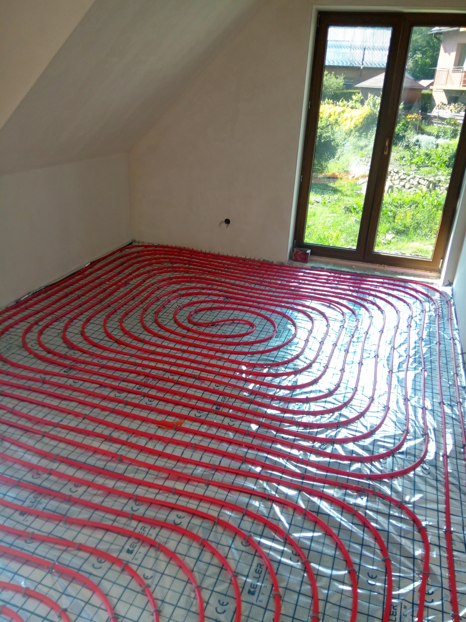 Czerwone rury ogrzewania podłogowego ułożone spiralnie na folii z siatką w jasnym pokoju na poddaszu z oknem.