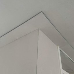 RenoKris KRZYSZTOF SZELĄG - Szary sufit i ściana z delikatną fakturą, połączone listwą, widoczny fragment okna.