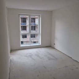 RenoKris KRZYSZTOF SZELĄG - Surowe wnętrze pokoju w nowym budownictwie z oknem na plac budowy, świeżo wykończone ściany w kolorze białym, surowa wylewka na podłodze.