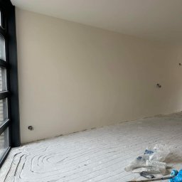 RenoKris KRZYSZTOF SZELĄG - Świeżo pomalowane wnętrze z widocznymi śladami po wałku na podłodze, zakrytej folią malarską, obok okna z czarną ramą i gładkimi ścianami w odcieniu beżu.