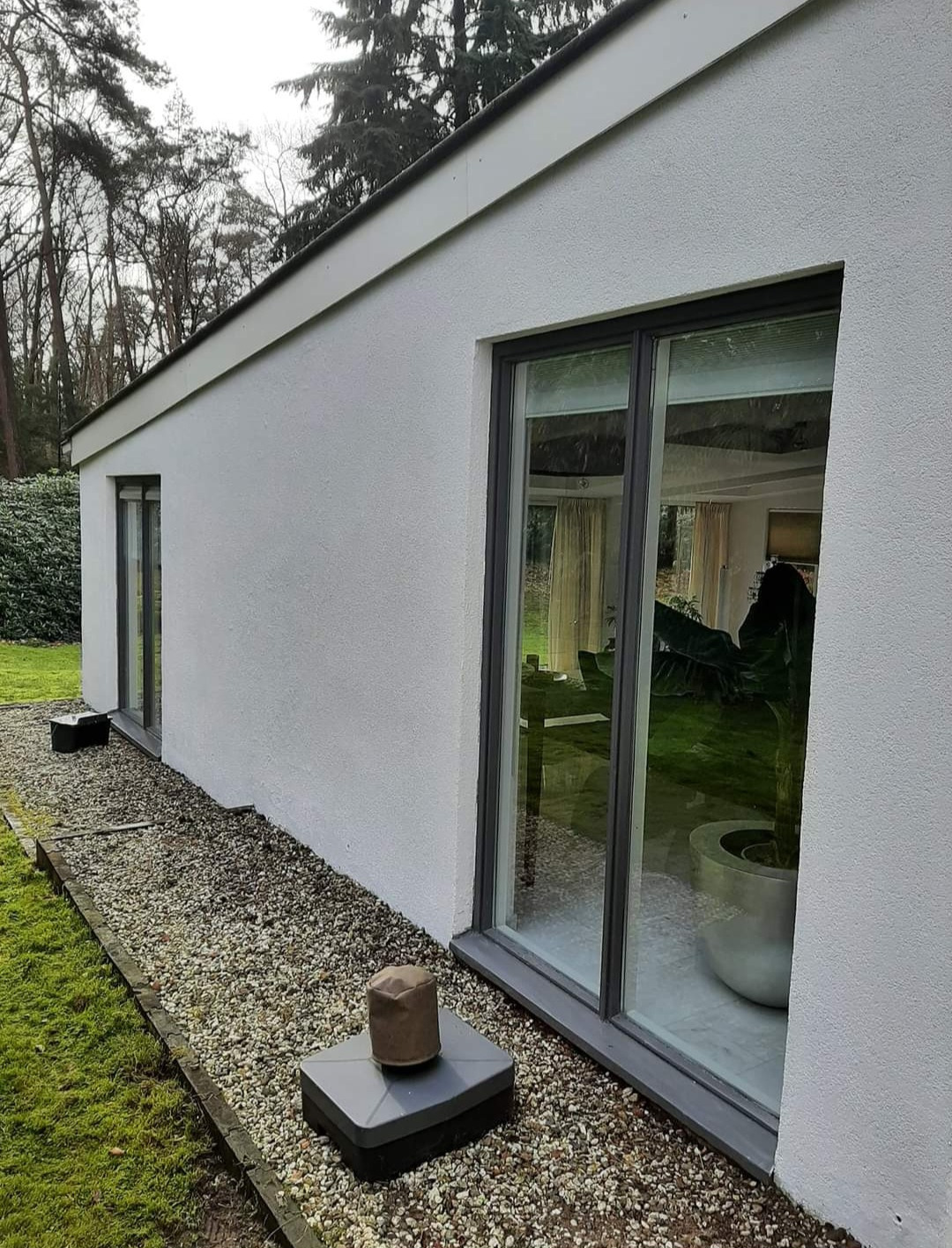 Biały budynek z szarymi ramami okiennymi, pokryty tynkiem o wyraźnej fakturze, z widocznymi elementami instalacji wentylacyjnej na zewnątrz i zielenią w tle.