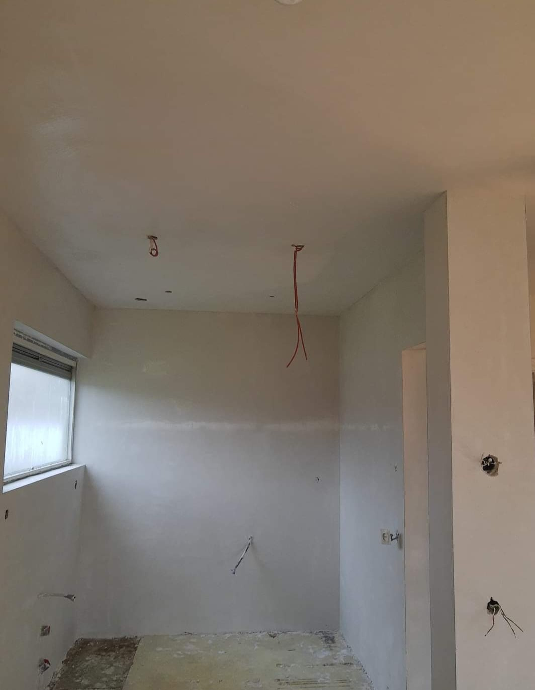 Wnętrze pomieszczenia w trakcie prac wykończeniowych: świeżo położone tynki na ścianach i suficie, wystające przewody elektryczne, fragment okna po lewej stronie, surowa podłoga.