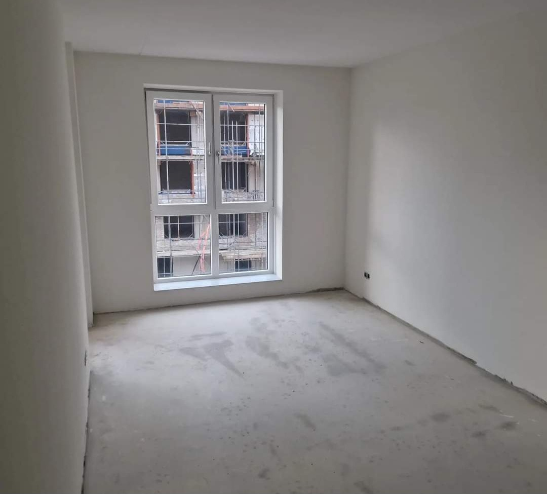 Surowe wnętrze pokoju w nowym budownictwie z oknem na plac budowy, świeżo wykończone ściany w kolorze białym, surowa wylewka na podłodze.