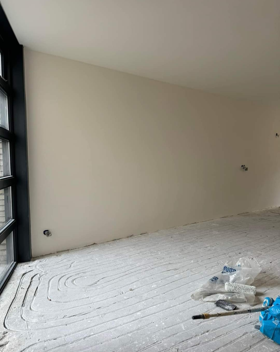 Świeżo pomalowane wnętrze z widocznymi śladami po wałku na podłodze, zakrytej folią malarską, obok okna z czarną ramą i gładkimi ścianami w odcieniu beżu.