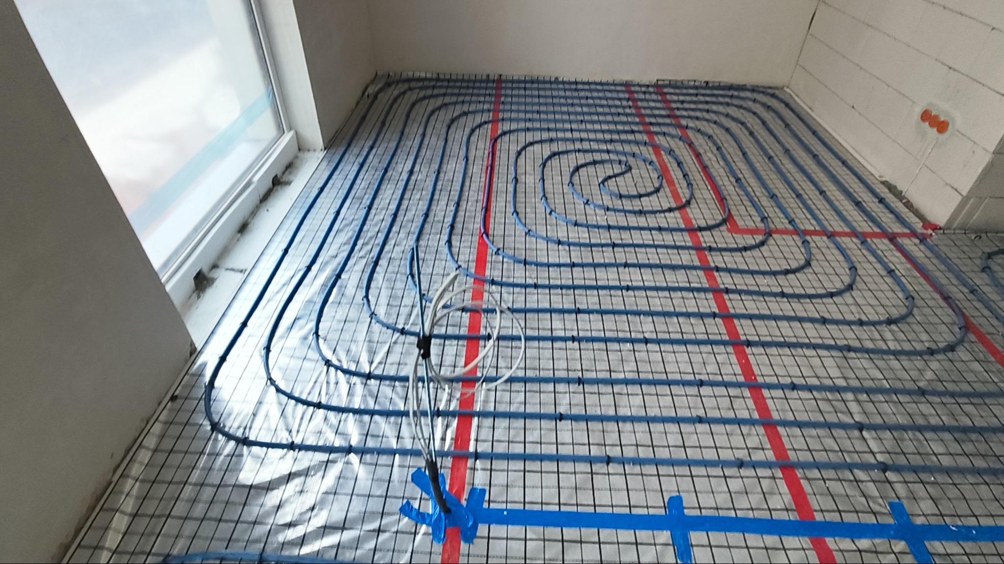Niebieskie rury ogrzewania podłogowego ułożone w spiralny wzór na srebrnej izolacji z siatką, z czerwonymi liniami montażowymi, w pomieszczeniu z oknem.