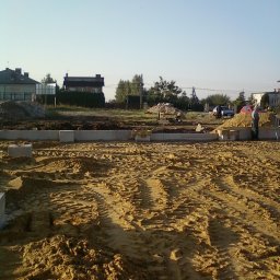 Piotr Dębek Mel-Bud firma budowlano-remontowa - Przygotowany teren pod fundamenty z widocznymi bloczkami betonowymi i śladami opon, w tle zabudowa jednorodzinna i osoba w jasnej odzieży przy pryzmie piasku.