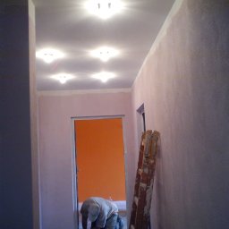 Piotr Dębek Mel-Bud firma budowlano-remontowa - Korytarz w trakcie prac wykończeniowych; ściany pokryte świeżo nałożoną gładzią w odcieniach różu, widoczne źródła światła w suficie i drzwi pomalowane na intensywny pomarańczowy kolor, drabina...