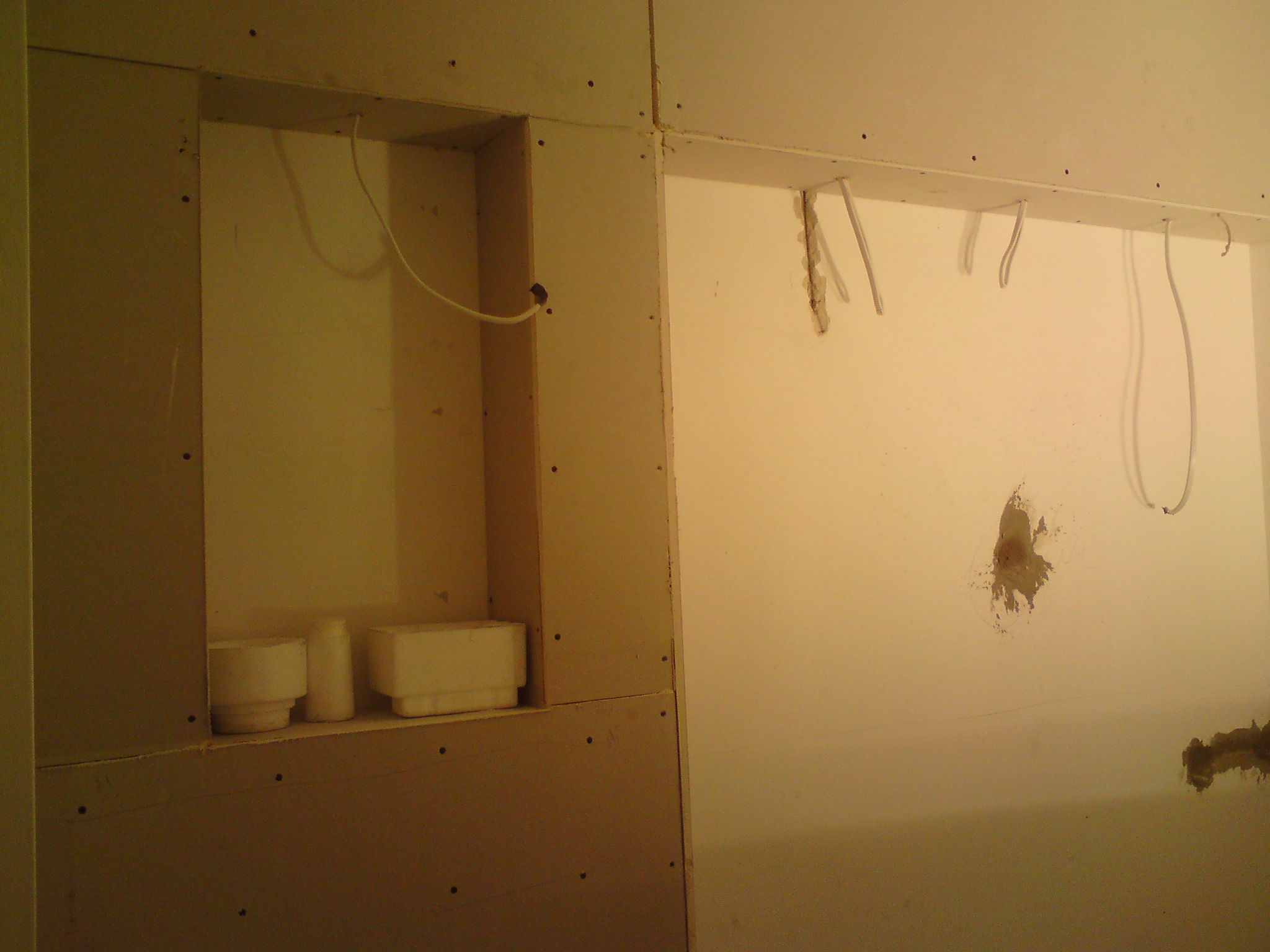 Ściana z płyt kartonowo-gipsowych w trakcie montażu, widoczne łączenia, niezabezpieczone przewody elektryczne wystające ze ściany oraz wnęka z elementami dekoracyjnymi.