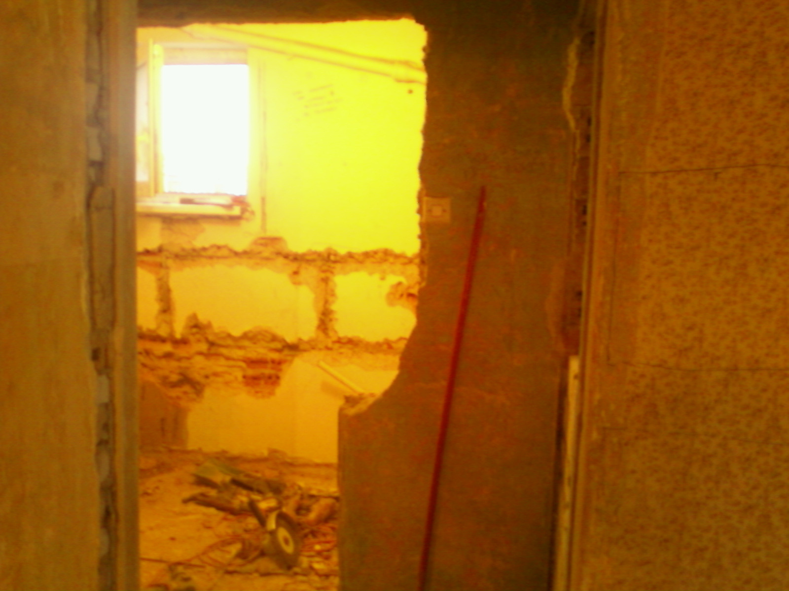 Wnętrze w trakcie demontażu ściany, widoczne odsłonięte cegły i gruz, narzędzia leżące na podłodze, intensywne żółte oświetlenie pomieszczenia.