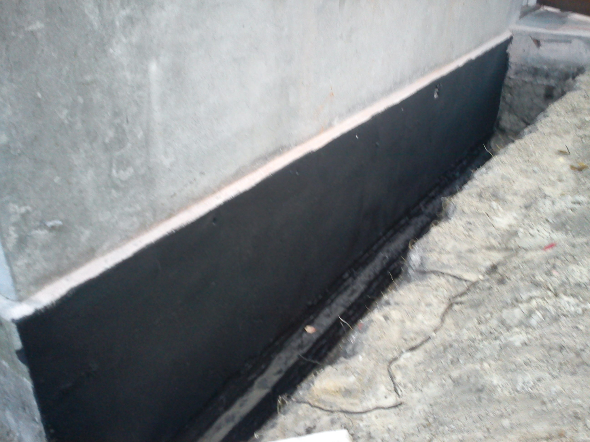 Hydroizolacja fundamentu, ciemna powłoka na betonowej ścianie, widoczne wykopane podłoże.