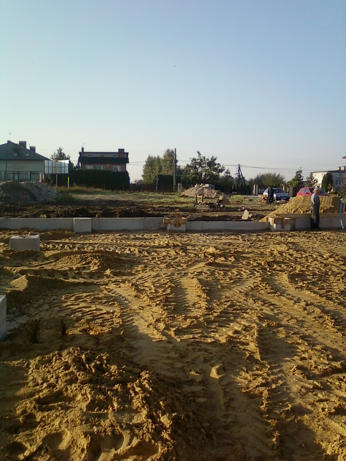 Przygotowany teren pod fundamenty z widocznymi bloczkami betonowymi i śladami opon, w tle zabudowa jednorodzinna i osoba w jasnej odzieży przy pryzmie piasku.