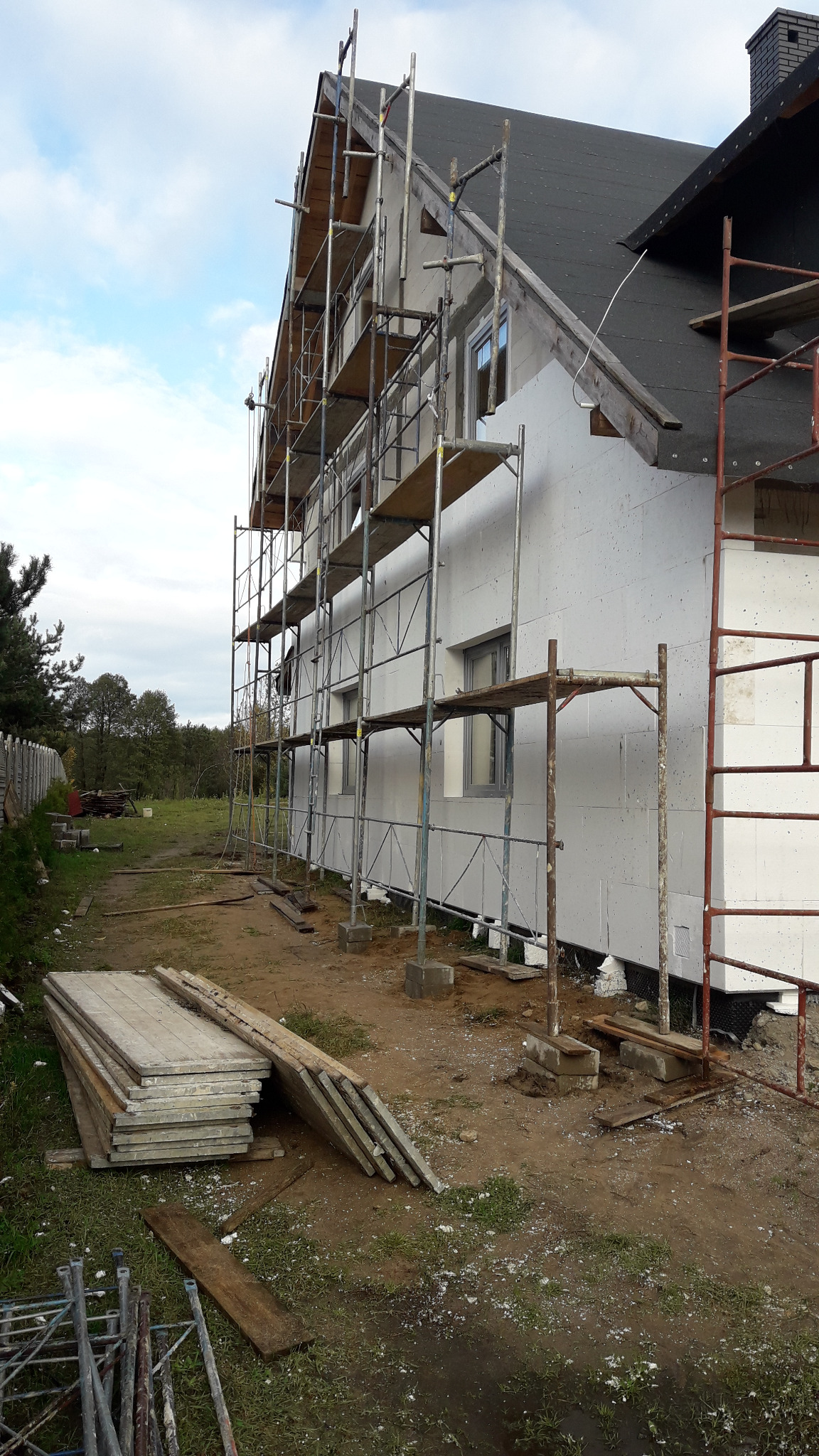 Dom w trakcie ocieplania styropianem, widoczne rusztowania, przygotowane płyty izolacyjne i fragment ogrodu.