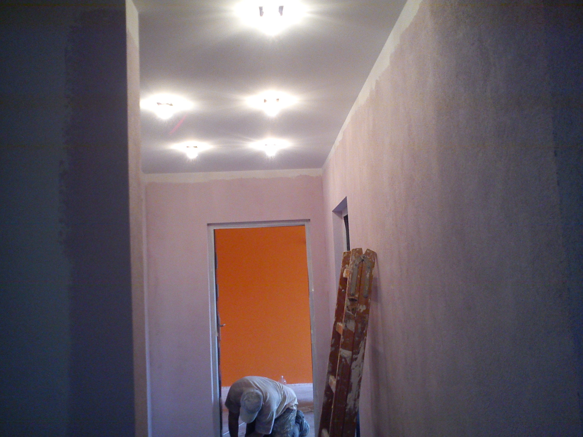 Korytarz w trakcie prac wykończeniowych; ściany pokryte świeżo nałożoną gładzią w odcieniach różu, widoczne źródła światła w suficie i drzwi pomalowane na intensywny pomarańczowy kolor, drabina...