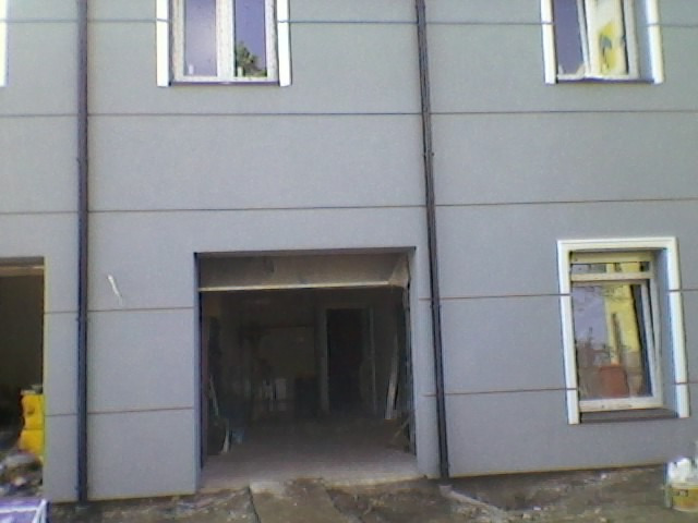 Szara fasada budynku z widocznymi oknami z białymi ramami i otwartym garażem z widocznym wnętrzem w trakcie prac wykończeniowych.