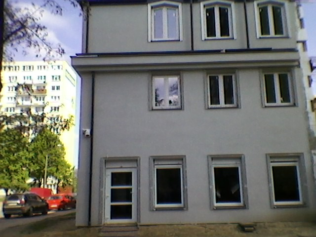 Fasada trzypiętrowego budynku mieszkalnego z szarym tynkiem, widoczne okna z białymi ramami, białe drzwi wejściowe, z boku zaparkowane samochody i inny budynek w tle.