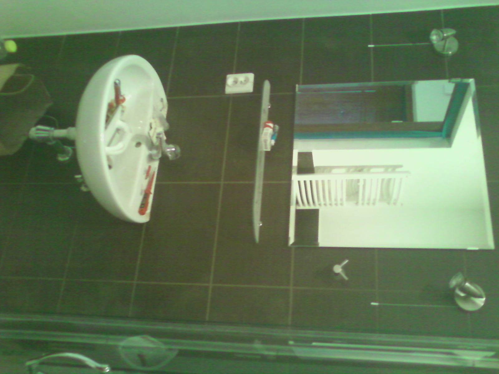 Umywalka ceramiczna z baterią, wisząca na ścianie z ciemnymi płytkami, obok lustro z widocznym grzejnikiem drabinkowym w tle, w łazience z widocznym gniazdkiem elektrycznym.