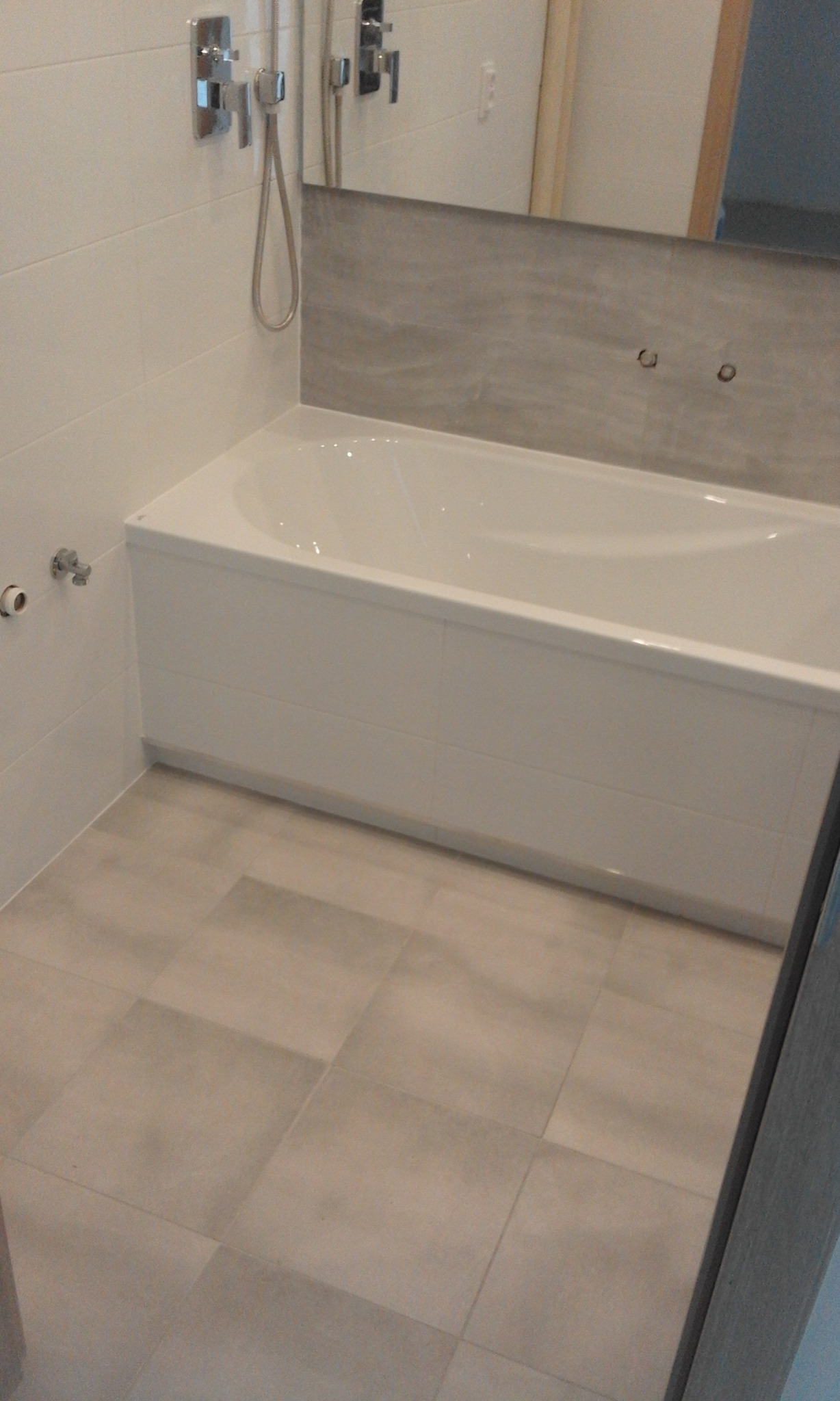 Biała wanna obudowana kafelkami, z chromowaną armaturą i szarą płytką nad wanną, na podłodze kwadratowe płytki w odcieniach beżu.