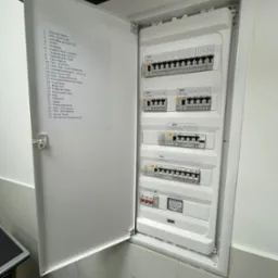 Otwarta skrzynka rozdzielcza z listwą zaciskową i wyłącznikami różnicowoprądowymi, opisana listą obwodów na wewnętrznej stronie drzwi, zamontowana na marmurowej ścianie.