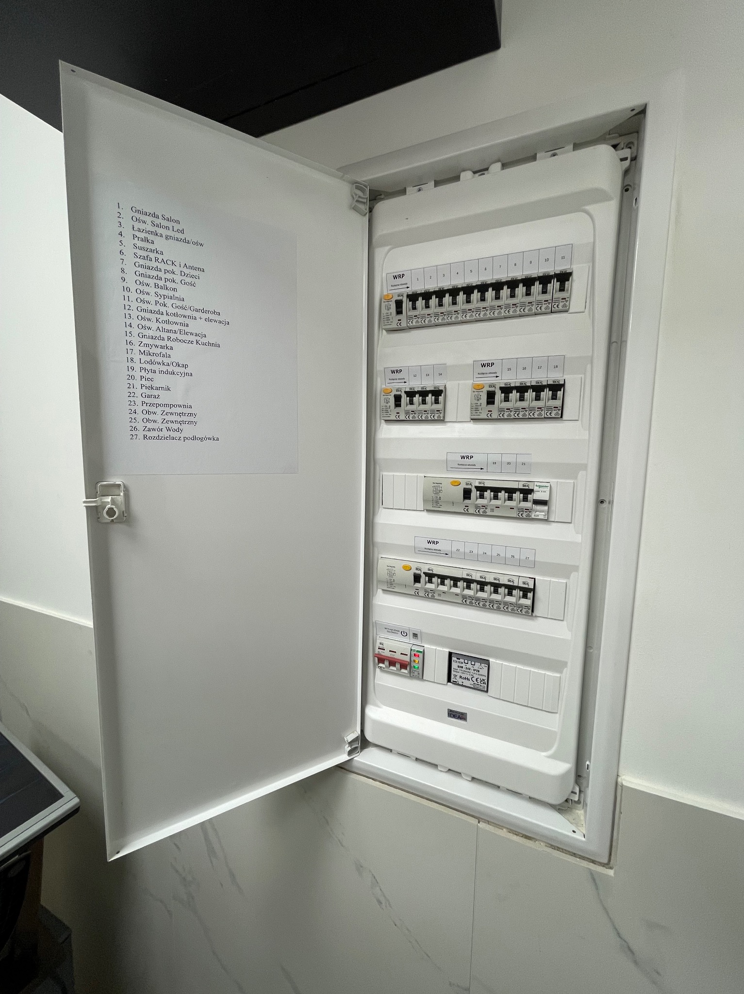 Otwarta skrzynka rozdzielcza z listwą zaciskową i wyłącznikami różnicowoprądowymi, opisana listą obwodów na wewnętrznej stronie drzwi, zamontowana na marmurowej ścianie.