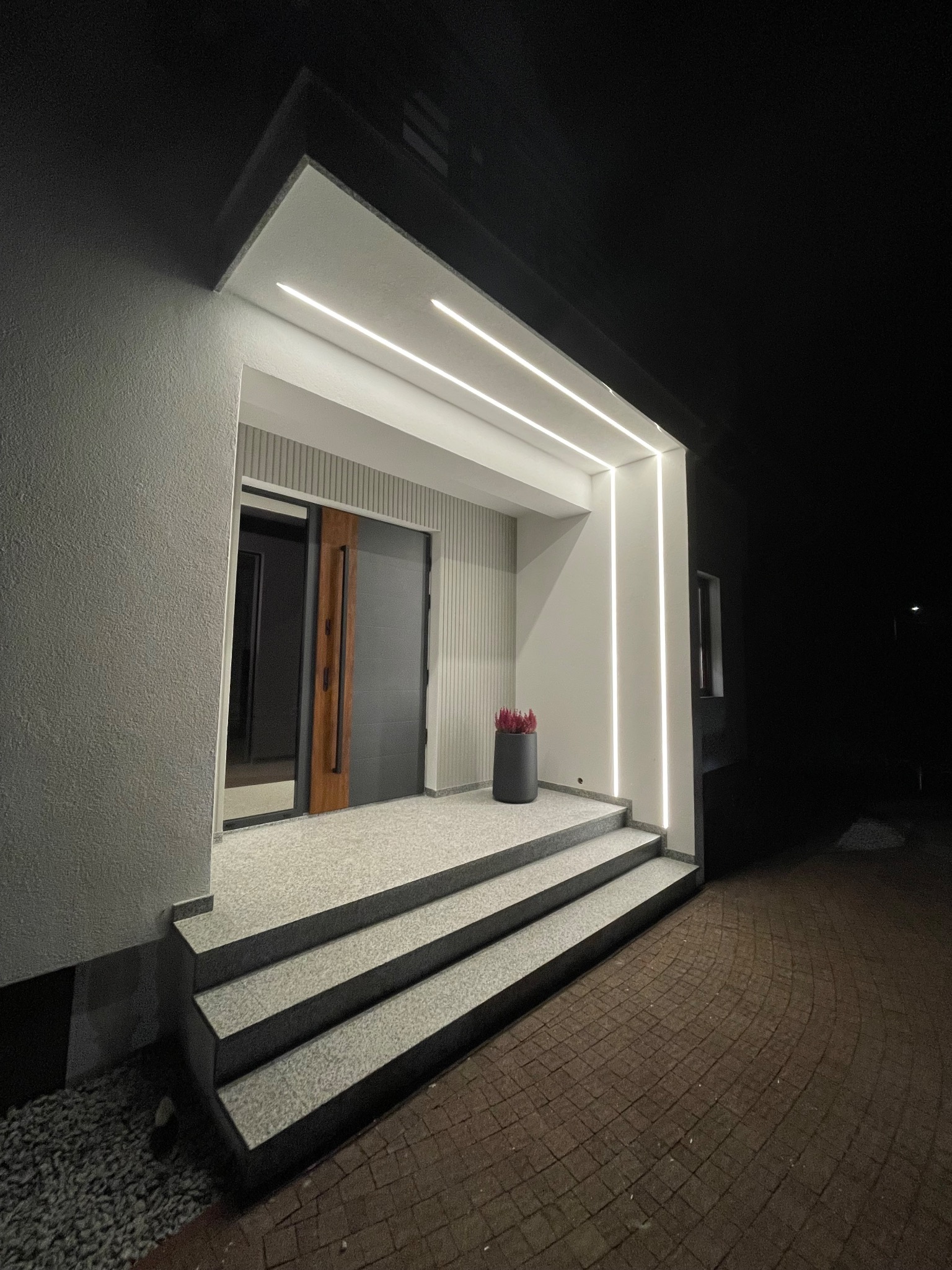 Nowoczesne wejście do domu z szarymi schodami, podświetleniem LED i ciemnymi drzwiami z drewnianym akcentem, widziane nocą.