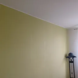 Ściana pokryta tapetą o delikatnej fakturze w odcieniach oliwkowej zieleni, widoczny fragment sufitu i narożnik pokoju z lampą podłogową, na której leży damski kapelusz.